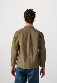 Olive green button-up shirt with long sleeves, featuring a back yoke and pleat detail, paired with blue trousers. Smooth texture.