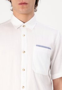 Camisa blanca de manga corta con frente abotonado, botones dorados y un bolsillo en el pecho de color azul claro. Textura suave y corte relajado.