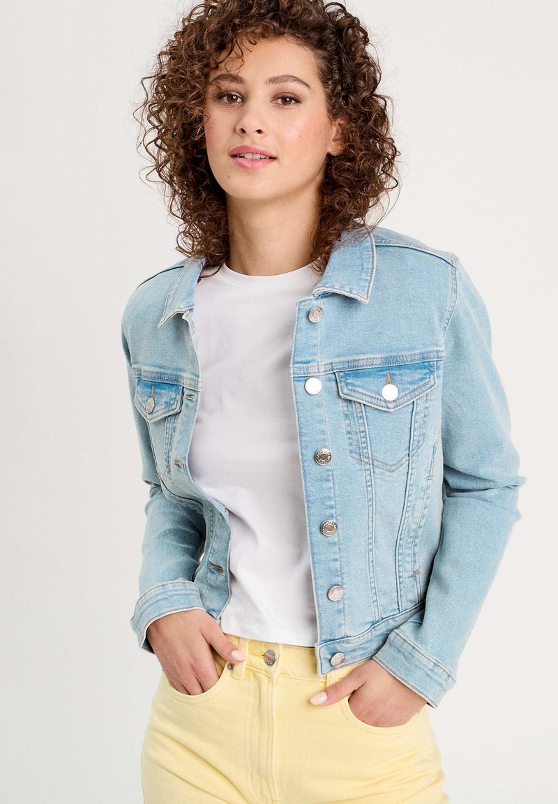Chaqueta de mezclilla azul claro con un corte clásico, cierre de botones, bolsillos en el pecho y costuras en contraste, llevada sobre una camiseta blanca y pantalones amarillos.