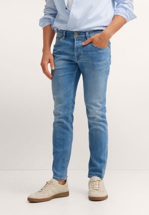 Person trägt hellblaue Slim-Fit-Jeans und beige Turnschuhe, steht mit einer Hand in der Tasche vor einem einfarbigen Hintergrund.