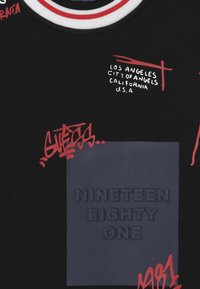 Musta lyhythihainen t-paita, jossa on punaiset ja valkoiset yksityiskohdat. Siinä on graffitityylinen teksti ja kuvioitu suorakulmainen kangasmerkki, jossa lukee "NINETEEN EIGHTY ONE."