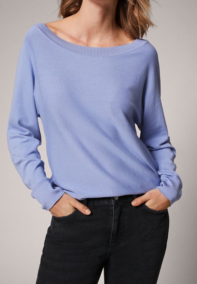 Pull violet clair à manches longues avec un large col et des poignets côtelés, associé à un jean foncé pour le contraste. Texture lisse, coupe décontractée.