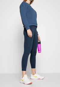 Granatowy sweatshirt o luźnym kroju z prążkowanymi mankietami, zestawiony z granatowymi legginsami. Białe trampki z żółtymi akcentami; trzyma różową butelkę na wodę.