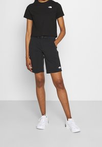 Svart kortärmad t-shirt med en liten vit logotyp, matchad med knälånga svarta shorts med bälte och sidofickor, bärs med vita sneakers.