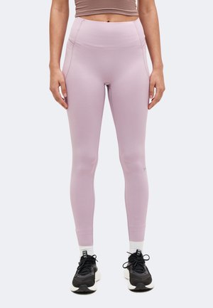 Personne portant des leggings violet clair, des chaussettes blanches et des baskets noires, debout les mains détendues le long du corps.