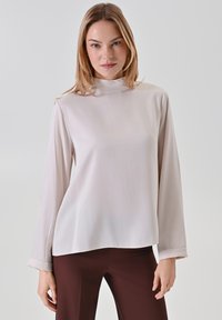 Helle beigefarbene Bluse mit langen Ärmeln und hohem Kragen. Sanfter, fließender Stoff mit lockerer Passform. Die Ärmel mit Bündchen reichen etwas über die Handgelenke.