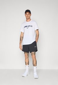 White Nike T-shirt with "Just Do It" text, paired with dark shorts. Worn with white sneakers and a dark cap, featuring intricate tattoos.