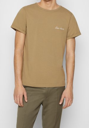 Mężczyzna w beżowej koszulce z podwiniętymi rękawami i białym napisem "zwolnij" na piersi, w połączeniu z oliwkowozielonymi spodniami.