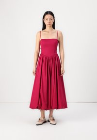 Vestido halter magenta con un cuerpo ajustado, falda acampanada y textura fruncida en la cintura. Lleva zapatos planos de color claro.