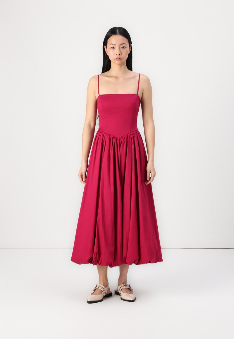 Vestido halter magenta con un cuerpo ajustado, falda acampanada y textura fruncida en la cintura. Lleva zapatos planos de color claro.