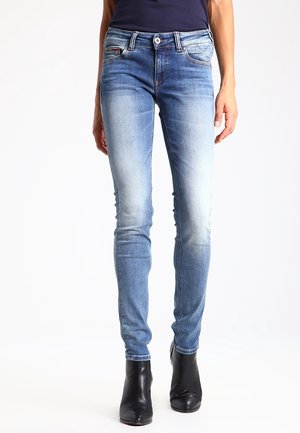 Donna con jeans skinny azzurri chiari e stivaletti neri, in piedi su sfondo bianco semplice.