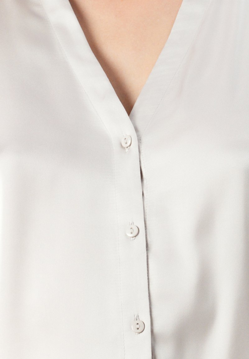 Blouse blanche en satin avec un décolleté en V et trois petits boutons blancs sur le devant, laissant entrevoir une partie du cou et du haut de la poitrine.