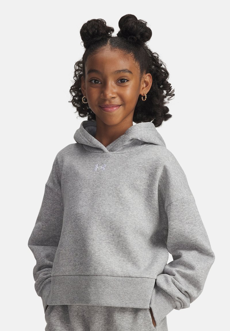 Girl with curly hair in two buns wears gray hoodie and matching pants, smiling with hands in pockets against plain background.