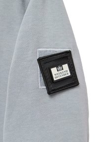Šedá mikina so štruktúrovanou čiernou nášivkou s nápisom "WEEKEND OFFENDER", ktorá obsahuje logo a je obklopená textúrovaným materiálom.