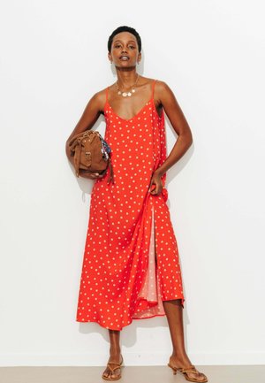 Femme en robe rouge à pois sans manches tenant un sac marron, portant des sandales et un collier en or, debout contre un mur blanc.