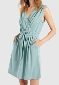 Une robe portefeuille en teinte bleu clair fabriquée dans un tissu doux, dotée d'un col en V profond, sans manches, d'une ceinture à la taille et de poches latérales.