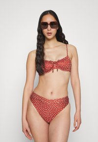 Biquíni vermelho com bolinhas brancas, com uma parte de cima tipo bandeau amarrada e parte de baixo de cintura alta. Tecido suave com um leve brilho e detalhes em dourado nas amarrações.