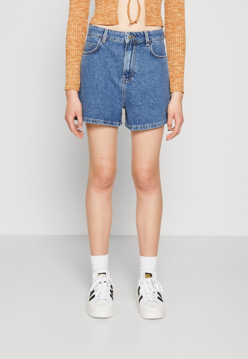 Personne portant un short en jean bleu taille haute, un crop top orange à manches longues, des chaussettes blanches montantes et des baskets blanches avec des rayures noires.