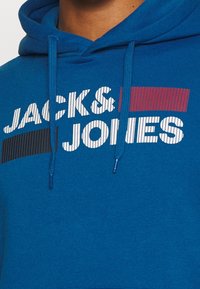 Modrá mikina s kapucí z měkké tkaniny s bílým, černým a červeným nápisem "JACK & JONES" s texturovanými pruhy, nastavitelnými šňůrkami a klokaní kapsou.