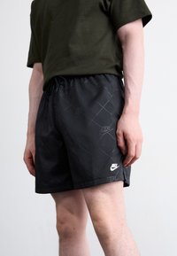 Sorte Nike shorts med et diamantmønster, der har en elastisk talje og snøre. Hvidt Nike-logo på kanten og siden. Glat stoftekstur.