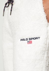 Vit, fluffig fleecejacka med marinblå dragsnören. Har broderad text "POLO SPORT" och en liten amerikansk flaggdetalj.