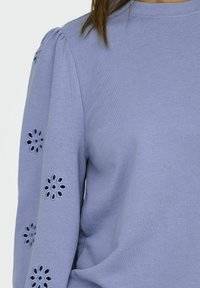 Ljuslila tröja med puffärmar och broderade blommönster i utskärningar längs ärmarna, bärs av en person med brunt hår.