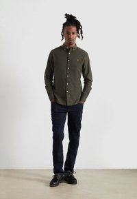 Camicia verde oliva con bottoni bianchi, vestibilità slim, indossata con jeans scuri e scarpe nere con lacci e suole tassellate.