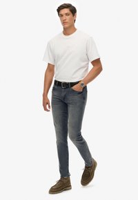 Camiseta blanca de manga corta, jeans ajustados gris oscuro, cinturón negro y zapatos de ante marrón. Presenta un reloj con una esfera azul en la muñeca.