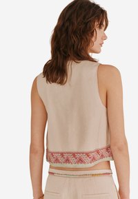 Top cropped sans manches beige en lin avec une broderie géométrique colorée le long de l'ourlet. Texture lisse avec des lignes épurées et un design minimal.