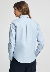 Kvinna med brunt hår som bär en ljusblå tröja med långa ärmar och svarta byxor, sedd bakifrån mot en enfärgad bakgrund.