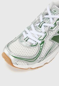 Chaussure de sport argentée avec tige en maille, accents verts, lacets blancs et semelle en caoutchouc gum. Remarquable pour son design léger et sa ventilation.
