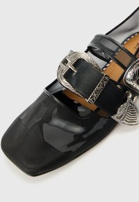 Schwarzer Schuh mit quadratischer Zehenform aus glänzendem Material mit einem Netzüberzug. Verfügt über eine silberne dekorative Schnalle und Lederakzente.