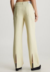 Pantalones de pierna ancha color amarillo pálido con bolsillos traseros y dobladillos con abertura. Presentan detalles de costura y un logo de CK en la cinturilla.