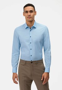 Chemise à manches longues bleu clair avec un col pointu, boutons noirs et une texture lisse, associée à un pantalon habillé marron.
