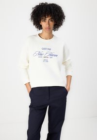 Sweatshirt ivoire avec texte graphique et détails d'emblème bleu marine, coupe décontractée, col rond et manches longues. Associé à un pantalon bleu marine foncé.