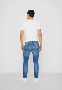 Jeans de mezclilla azul de corte ajustado con una cintura media, que cuentan con dos bolsillos traseros y un pequeño parche de cuero tan con la marca. Combinados con una camiseta blanca ajustada.