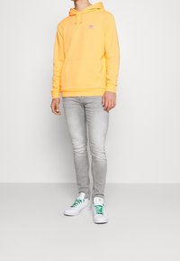 Gul hoodie med framficka och lila logotyp, kombinerad med grå slim-fit jeans och vita sneakers med gröna snören.