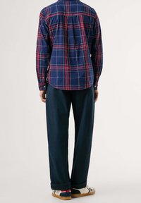 Osoba ubrana w koszulę z długim rękawem w granatowo-czerwono-białą krateczkę, ciemnoniebieskie spodnie i białe trampki, stoi tyłem.