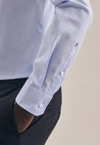 Lichtblauw overhemdbloes met een gladde stof, witte knopen en een klein donker logo op de manchet. De mouw is iets getailleerd.