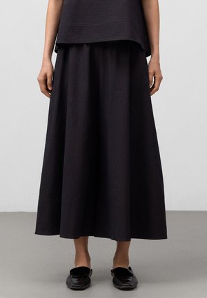 Gonna nera, strutturata, lunga fino al ginocchio con un design ampio e fluido. Abbinata a semplici scarpe nere senza lacci. Estetica moderna e minimalista.