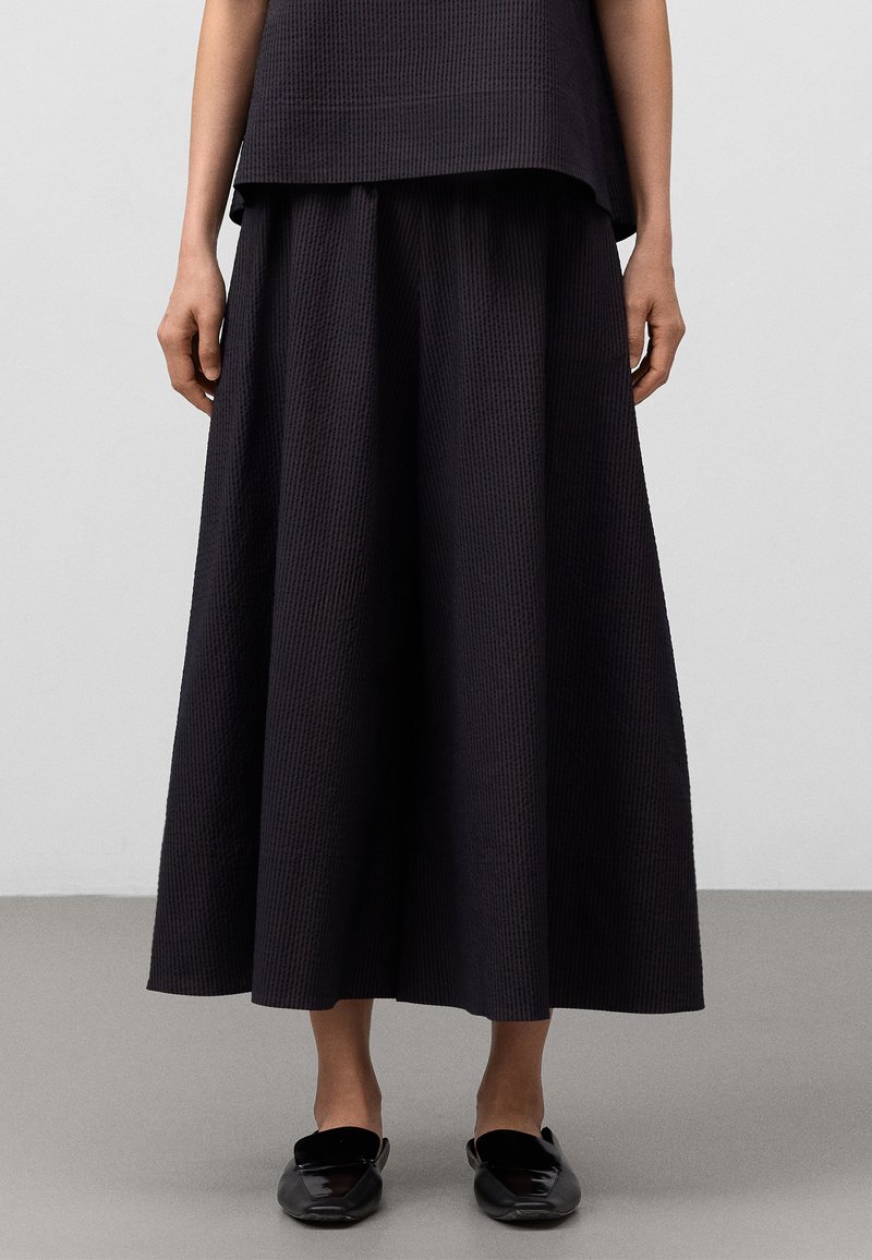 Schwarzer, strukturierter Rock in Knielänge mit weitem, fließendem Design. Kombiniert mit schlichten schwarzen Slippers. Moderner, minimalistischer Stil.