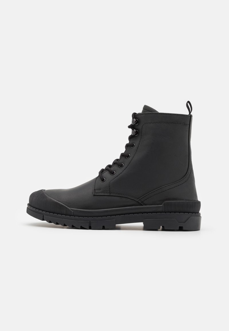 Botte de combat noire à lacets avec une semelle robuste, un bout arrondi, un col rembourré et une languette sur un fond clair uni, vue de côté extérieur.
