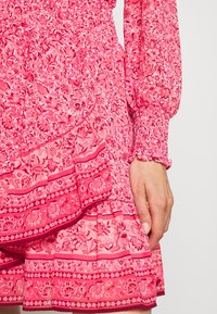 Rosa blommig klänning med volangerade lager och smockade manschetter. Tyget har intrikata blommönster i olika nyanser av rosa.