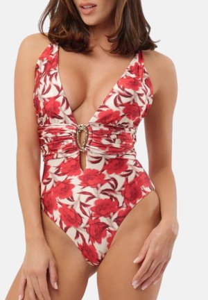 Donna che indossa un costume intero bianco con stampa floreale rossa e un dettaglio arricciato centrale con una fibbia ovale decorativa.