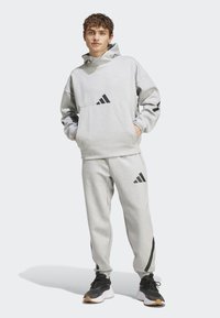 Sivá mikina s kangaroo vreckom a čiernym logom s tromi pruhmi, v kombinácii s nohavicami na doma s tapered strihom, ktoré majú čierne akcenty a logo.