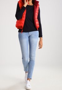 Gilet imbottito rosso con collo alto, indossato sopra una maglietta nera. Jeans fitted blu chiaro e sneakers bianche completano il look.