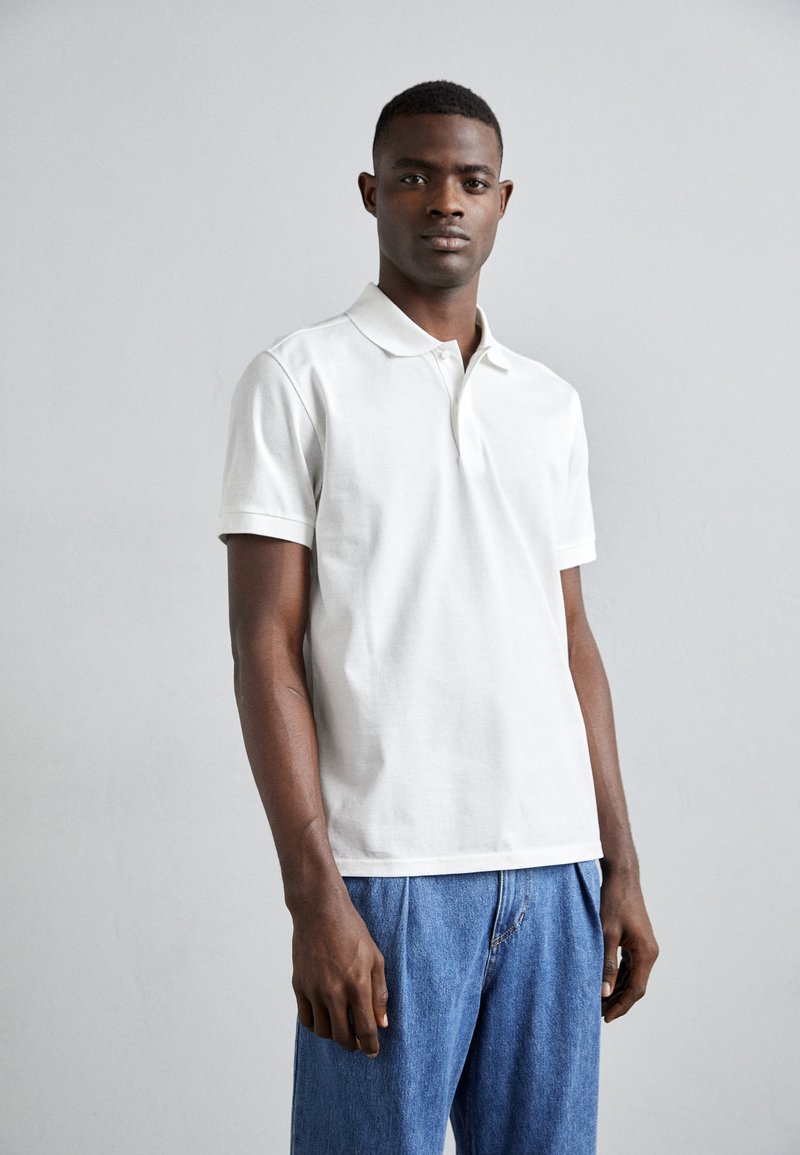 Camisa polo de algodón blanca con cuello clásico y mangas cortas, combinada con pantalones de mezclilla azul de corte relajado y costuras visibles.