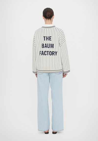 Veste oversize rayée blanche avec lettrage bleu marine "THE BAUM FACTORY" dans le dos, associée à un jean large bleu clair.