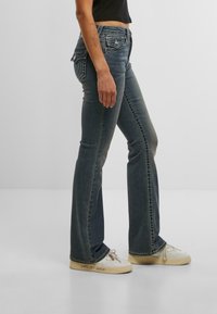 Zavihani traper hlače v temno modri barvi s kontrastnim šivanjem, vsebujejo zadnje žepke in rahlo zbledel videz. Ujemajo se z belimi supergami.