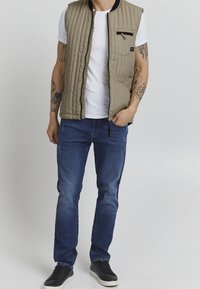 Gilet trapuntato beige con una tasca zip, abbinato a una t-shirt bianca e jeans blu. Sneakers nere completano l'outfit.